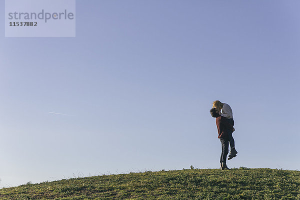 Junges Paar  das sich auf einem Hügel vor blauem Himmel küsst.