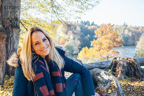 Lächelnde Frau genießt den Herbst im Wald auf dem Baumstamm