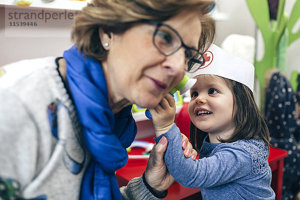 Porträt eines kleinen Mädchens  das das Ohr seiner Großmutter mit medizinischem Spielzeug untersucht.