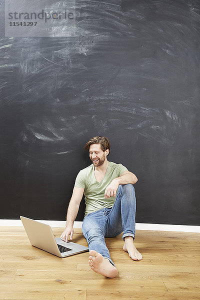 Porträt eines lächelnden jungen Mannes  der mit einem Laptop vor der Tafel auf dem Boden sitzt.