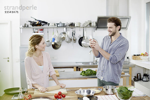 Mann fotografiert lächelnde Frau bei der Zubereitung von Pizzateig in der Küche.