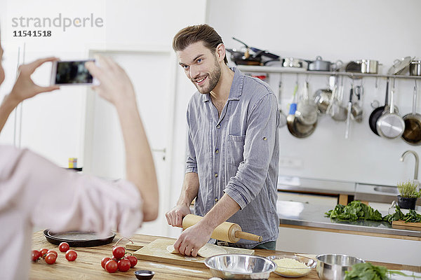 Frau macht Handyfoto von lächelndem Mann bei der Teigzubereitung in der Küche