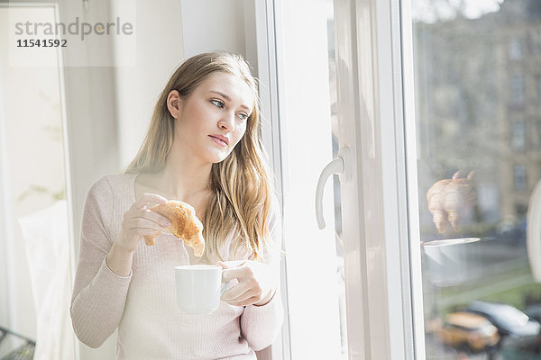 Porträt der jungen Frau mit Croissant und Tasse Kaffee durchs Fenster schauend