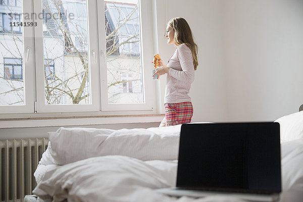 Junge Frau mit Croissant und Tasse Kaffee stehend im Schlafzimmer mit Blick durchs Fenster