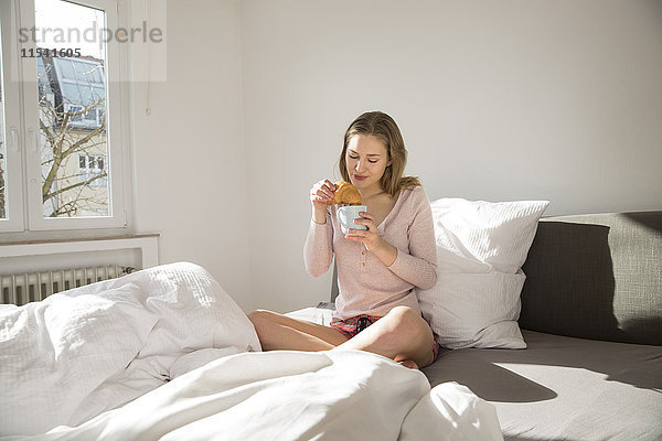 Junge Frau auf dem Bett sitzend mit Kaffee und Croissant