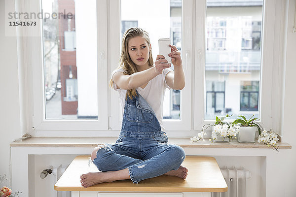 Junge Frau  die zu Hause auf einem Tisch sitzt und Selfie mit dem Smartphone nimmt