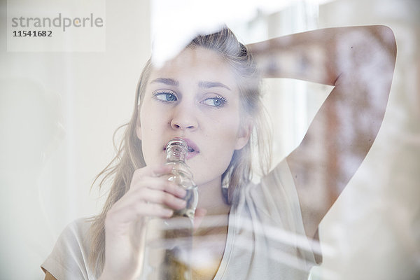 Porträt einer jungen Frau  die beim Trinken durch die Fensterscheibe schaut