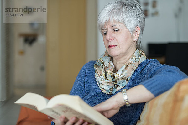 Eine Seniorin sitzt am Fenster auf der Couch und liest ein Buch. Verfügbares Licht. Grevenbroich  Nordrhein-Westfalen  Deutschland