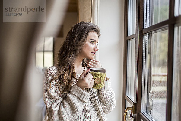 Junge Frau zu Hause mit Blick aus dem Fenster bei einer Tasse Kaffee