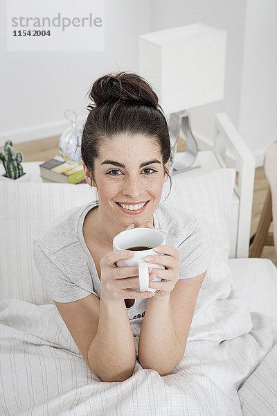 Porträt einer lächelnden jungen Frau mit einer Tasse Kaffee im Bett