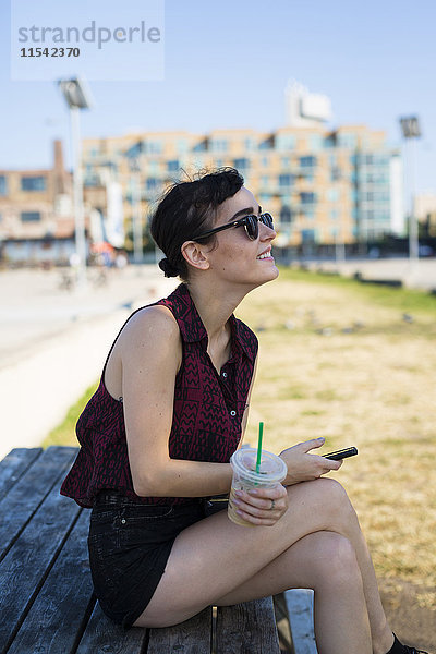 USA  New York City  Brooklyn  junge Frau sitzt auf einer Bank und hält Smartphone und Plastikbecher.