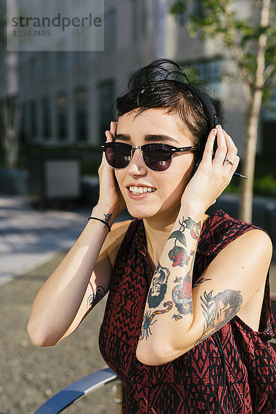 Portrait der tätowierten jungen Frau mit Kopfhörer und Sonnenbrille