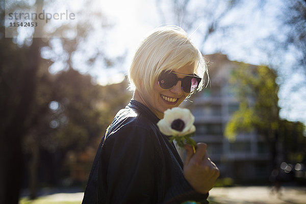Porträt einer lächelnden blonden Frau mit einer Sonnenbrille  die eine Blume hält.