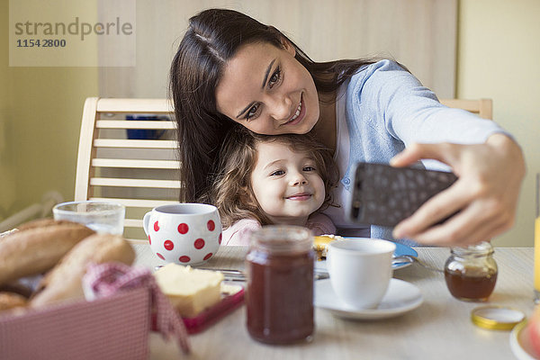 Porträt der Mutter und ihrer kleinen Tochter  die einen Selfie am Frühstückstisch nehmen