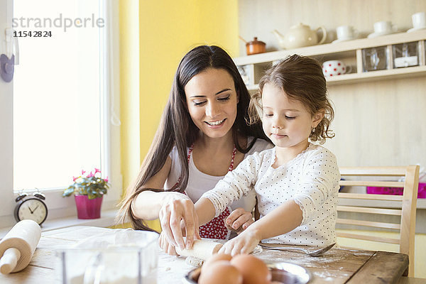 Porträt eines kleinen Mädchens und ihrer Mutter  die zusammen auf dem Küchentisch Teig ausrollen.