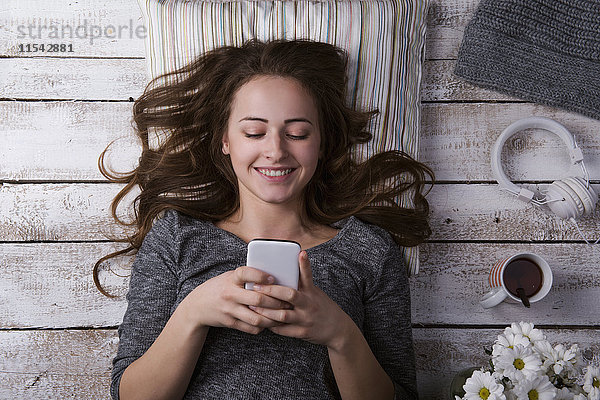 Porträt einer lächelnden jungen Frau  die auf dem Boden liegt und ihr Smartphone ansieht.