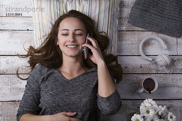 Porträt einer lächelnden jungen Frau  die auf dem Boden liegt und mit ihrem Smartphone telefoniert.