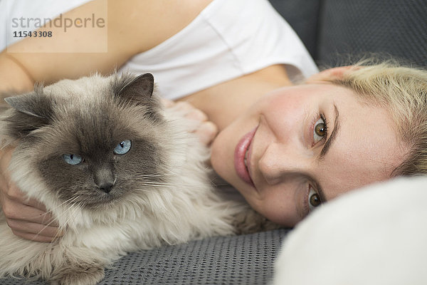 Porträt einer lächelnden jungen Frau  die mit ihrer Katze auf der Couch liegt.