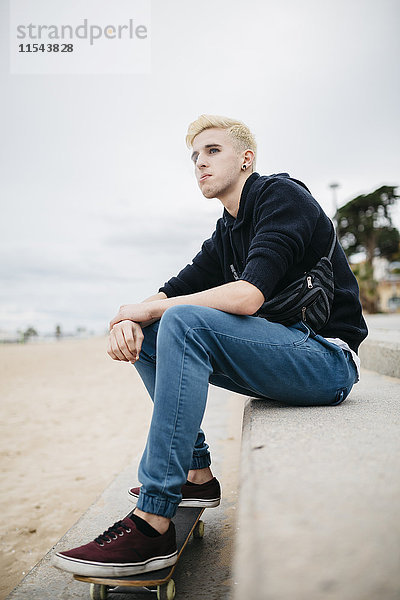 Spanien  Torredembarra  rauchender junger Skateboarder auf einer Wand am Strand sitzend