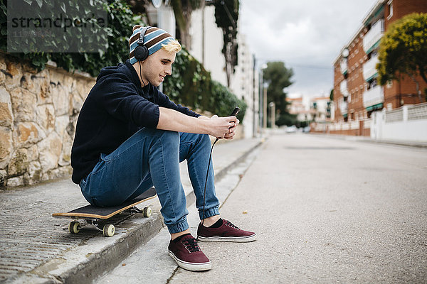 Spanien  Torredembarra  lächelnder junger Mann sitzt auf seinem Skateboard und hört mit Kopfhörern Musik.