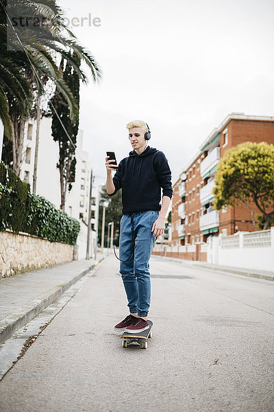 Spanien  Torredembarra  junger Mann mit Kopfhörern auf dem Skateboard und Blick auf sein Smartphone