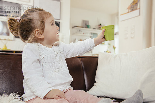 Kleines Mädchen hält einen Plastikbecher hoch  während es auf der Couch sitzt.
