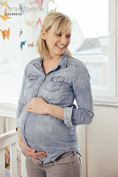 Porträt einer schwangeren Frau  die ihren Bauch im Kinderzimmer hält.