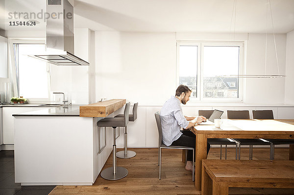 Mann sitzt am Tisch in seiner offenen Küche mit Laptop