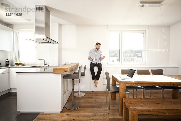 Mann mit Kaffeetasse auf Sideboard in seiner offenen Küche mit Blick auf Smartphone