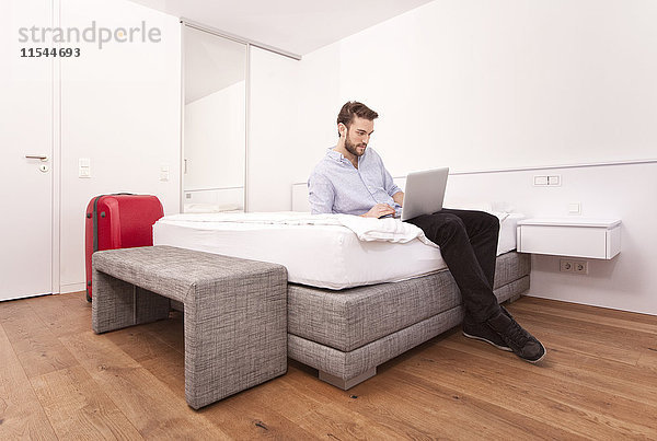 Junger Mann sitzend auf einem Hotelbett mit Laptop