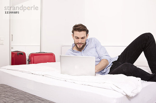 Junger Mann auf einem Hotelbett liegend mit Laptop