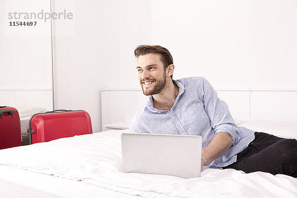 Porträt des lächelnden jungen Mannes auf Hotelbett mit Laptop