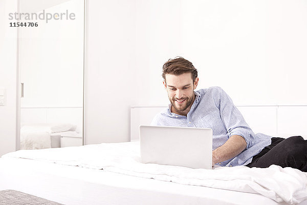 Lächelnder junger Mann auf einem Hotelbett liegend mit Laptop