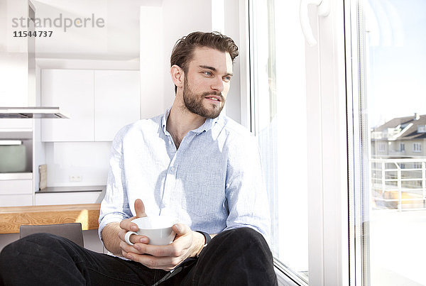 Junger Mann mit Kaffeetasse auf Fensterbank sitzend durchs Fenster schauend