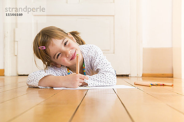 Porträt eines lächelnden Mädchens auf Holzboden mit Buntstiften und Papierbogen