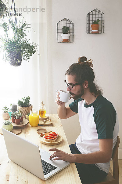Junger Mann mit Laptop beim Frühstücken
