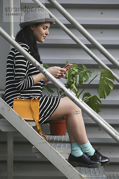 Lächelnde junge Frau sitzt auf der Treppe und schaut auf ihr Smartphone.