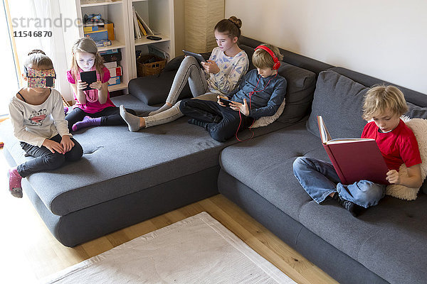 Vier Kinder auf einer Couch mit verschiedenen digitalen Geräten  während ein Junge ein Buch liest.