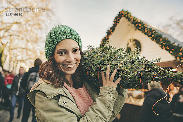 Glückliche Frau mit einem eingewickelten Baum  die über den Weihnachtsmarkt läuft.