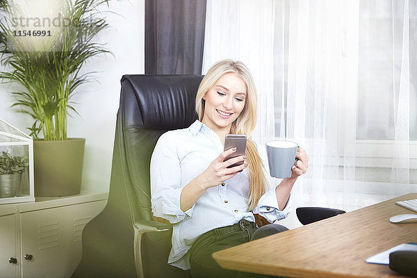 Porträt einer lächelnden blonden Frau am Schreibtisch mit einer Tasse Kaffee auf ihrem Smartphone.