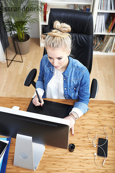 Blonde Frau sitzt am Schreibtisch und arbeitet mit einem Grafiktablett.