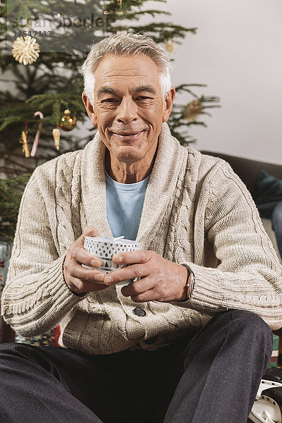 Porträt eines älteren Mannes mit Weihnachtsgeschenk vor dem Baum sitzend