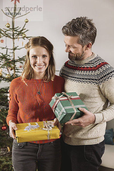 Paar mit Weihnachtsgeschenken vor dem Baum stehend