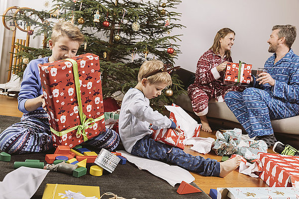 Kleiner Junge beim Auspacken eines Weihnachtsgeschenks  Eltern sitzen auf der Couch im Hintergrund