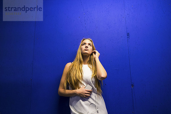 Porträt einer blonden Frau  die sich an die blaue Wand lehnt und mit Kopfhörern Musik hört.