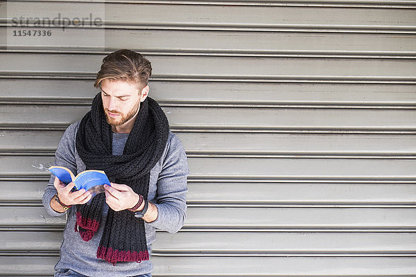Mann mit Schal steht vor dem Rollladen und liest ein Buch.