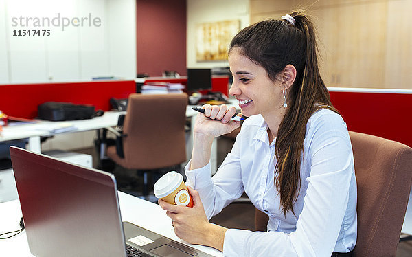 Lächelnde junge Geschäftsfrau mit Kaffee zum Sitzen am Schreibtisch