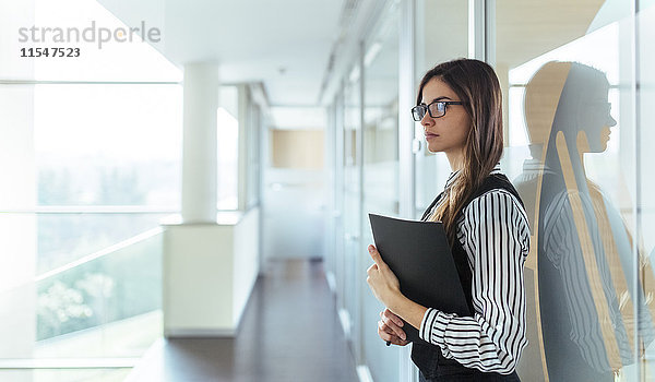 Junge Frau mit schwarzer Akte am Flur eines Büros stehend