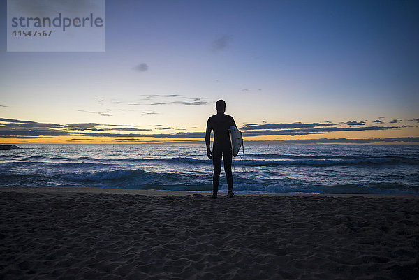 Rückansicht des am Strand stehenden Surfers bei Sonnenaufgang