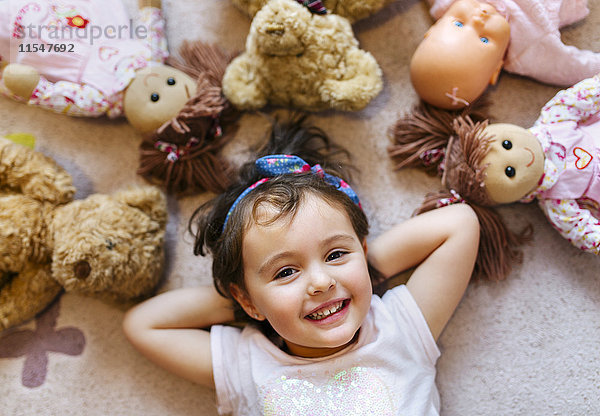 Porträt eines lächelnden kleinen Mädchens auf dem Boden liegend mit Teddys und Puppen um sie herum.
